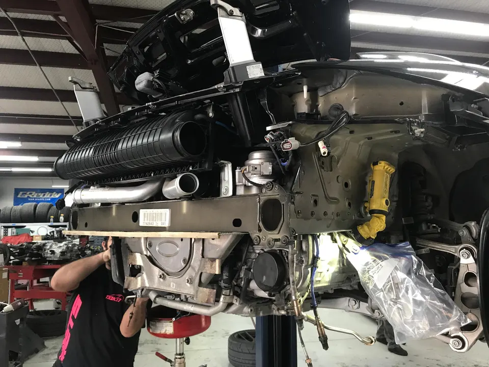Mechanic working on the front end of a Porsche 911 on a lift, with intercooler and boost piping visible.