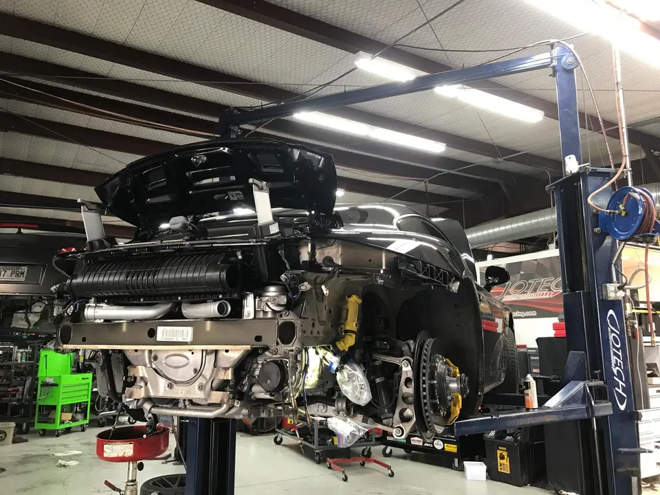 A black Porsche 911 on a lift in a performance shop, undergoing work with its rear bumper removed, revealing the intercooler and suspension.