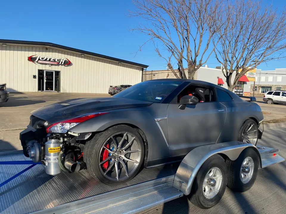 A gray Nissan GT-R R35 with its front bumper removed, revealing a large turbocharger, on a trailer in front of Jotech Motorsports.