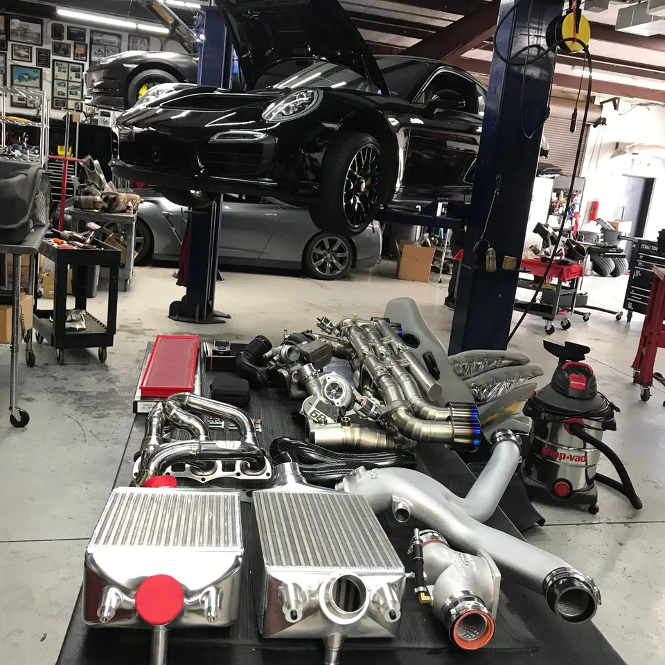 A black Porsche on a lift in a performance shop with a table full of turbochargers, intercoolers, and exhaust parts.