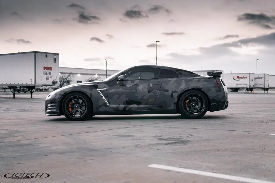 Side profile of a custom Nissan GT-R R35 with a dark camouflage wrap and aftermarket wheels in an industrial parking lot.