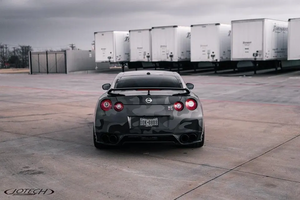 Rear view of a Nissan GT-R R35 with a dark camouflage wrap and aftermarket spoiler, parked in an industrial setting.