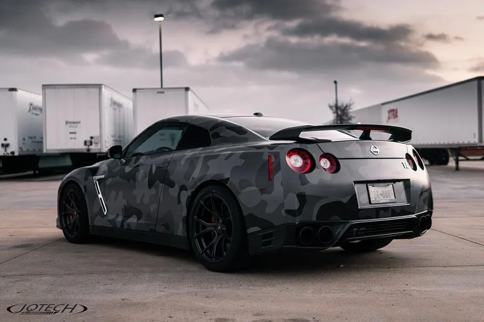 A custom Nissan GT-R R35 with a grey camouflage wrap, aftermarket black wheels, and a large rear wing, parked outdoors.