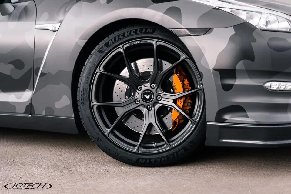 A close-up of a Nissan GT-R with a grey camouflage wrap, featuring aftermarket dark grey wheels, Michelin tires, and orange performance brake calipers.