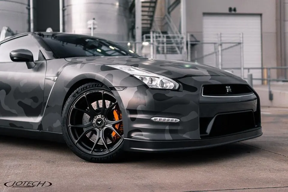 A custom Nissan GT-R R35 with a grey camouflage wrap, black Vossen wheels, and orange brake calipers parked outdoors.