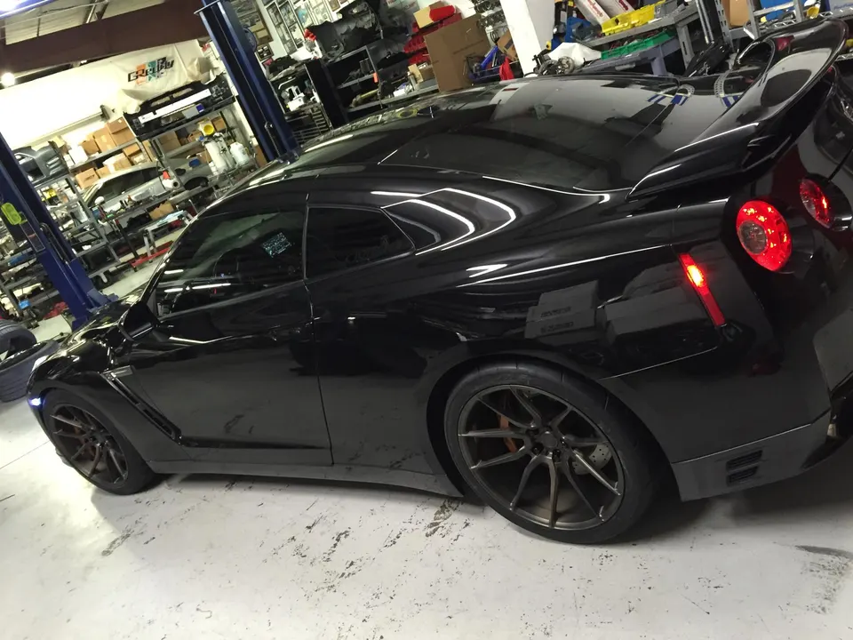 A black Nissan GT-R with aftermarket wheels parked in a performance tuning shop, showing the rear and side.