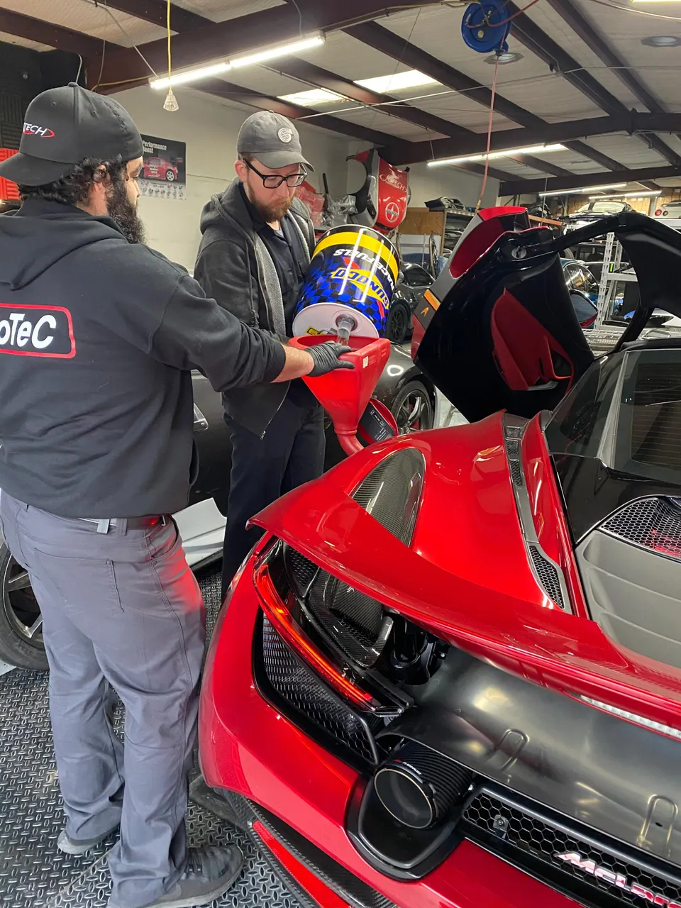 Two mechanics fueling a red McLaren supercar with race fuel from a drum in a performance tuning shop.