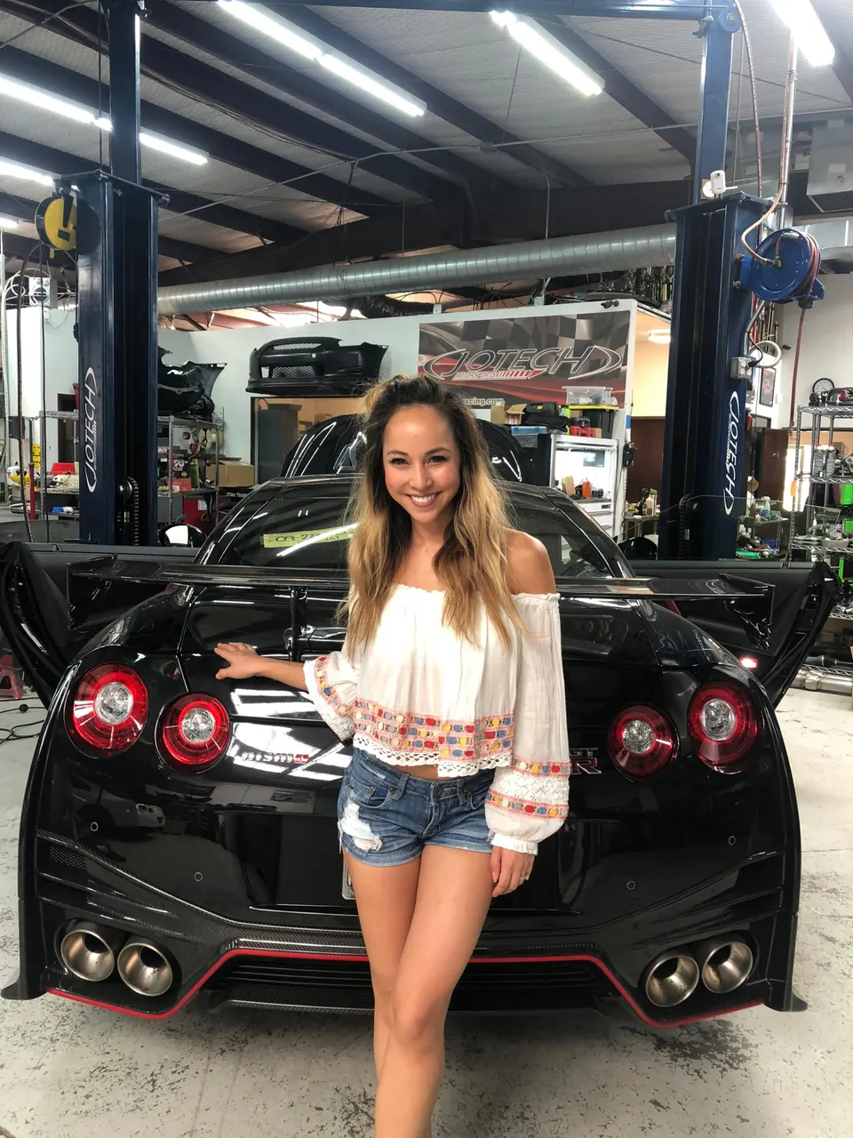 Woman posing with a black Nissan GT-R in a performance tuning shop, showcasing its aftermarket exhaust and carbon fiber diffuser.