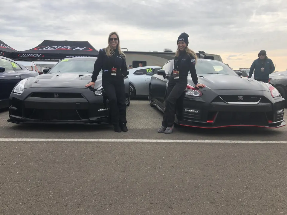 Two women in racing suits pose with black Nissan GT-R R35s at a Jotech Motorsports track event.