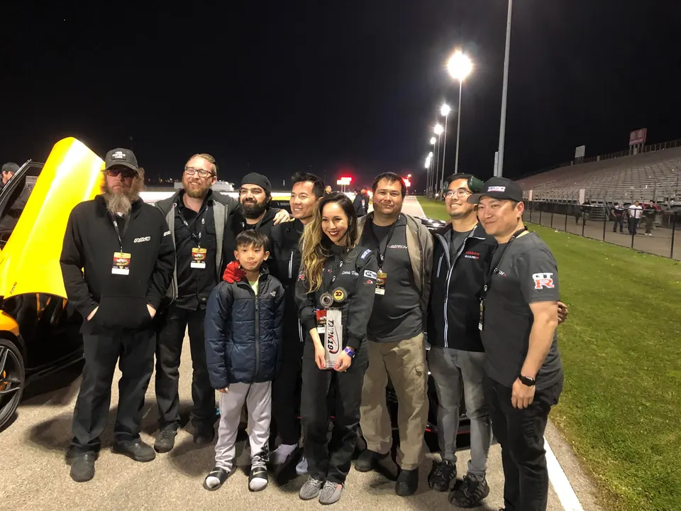 A group of people, including a child, pose for a photo next to a yellow car at a racetrack at night.