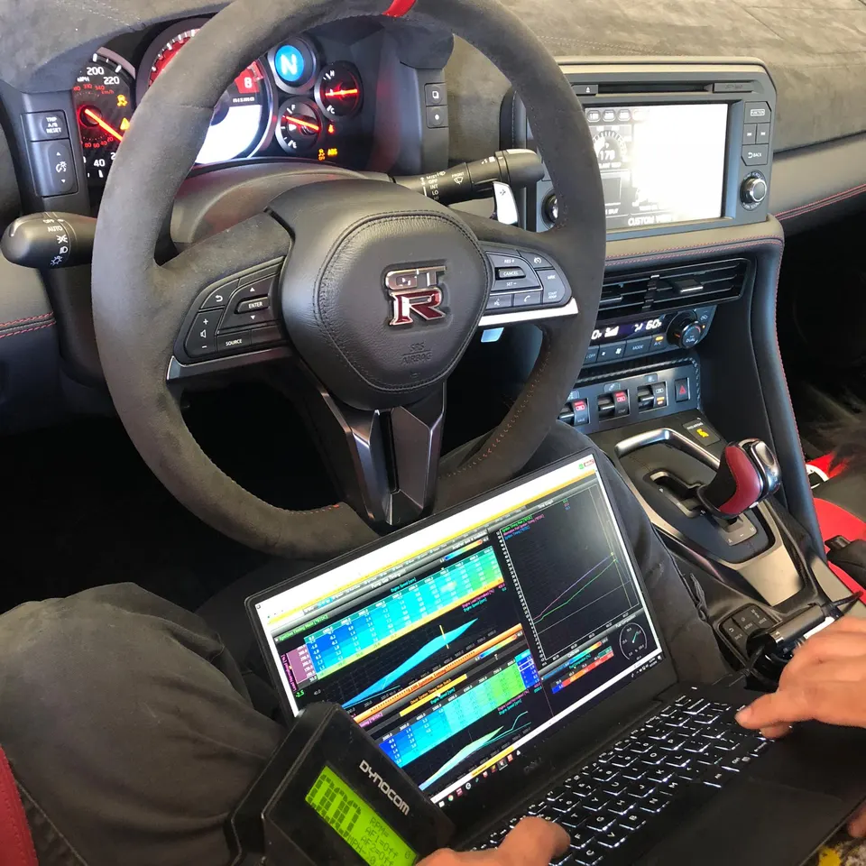 Tuner working on a Nissan GT-R interior with a laptop and Dynocom device, displaying tuning data and graphs.
