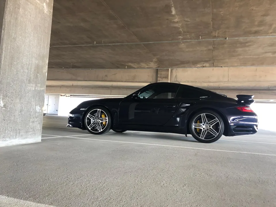 Dark blue Porsche 911 Turbo parked in a concrete parking garage, showcasing its side profile and rear wing.
