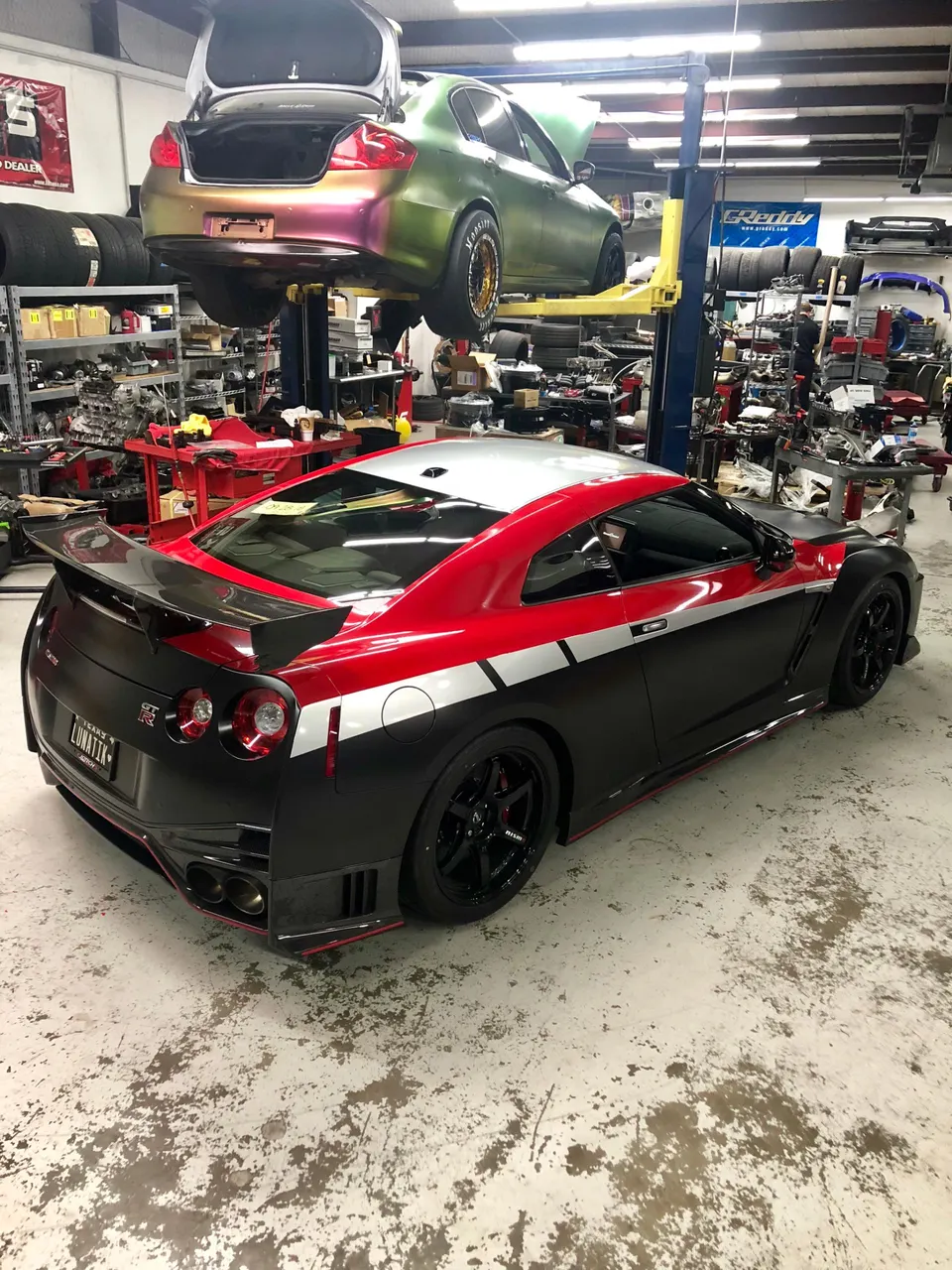 A custom black and red Nissan GT-R R35 with a widebody kit in a performance tuning shop with another car on a lift.