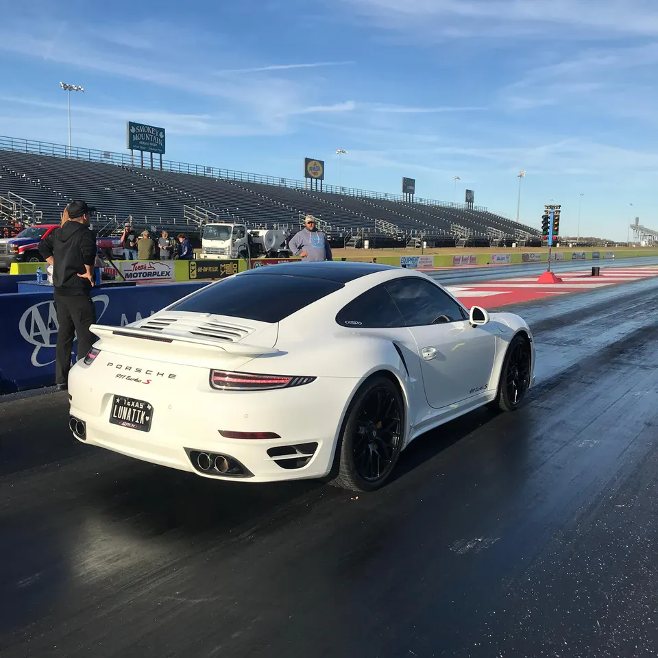 White Porsche 911 Turbo S with black wheels on a drag strip, with grandstands in the background.