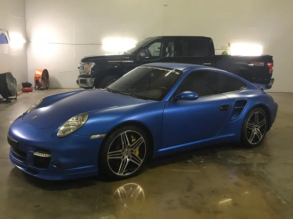 A stunning blue matte-wrapped Porsche 911 sports car with custom wheels, parked in a brightly lit garage.
