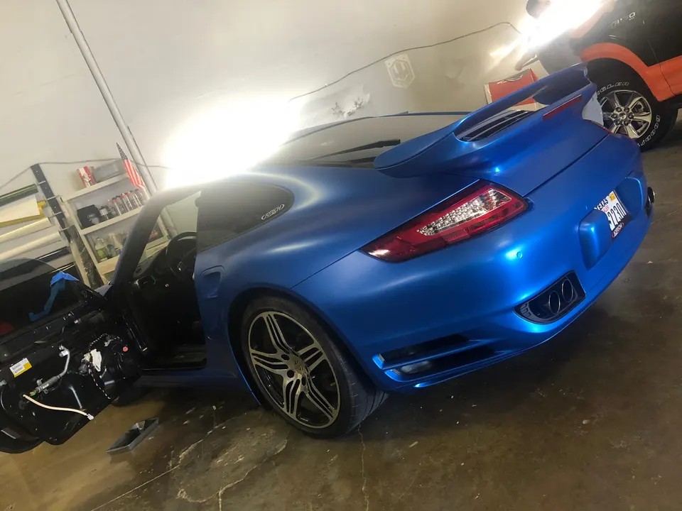 Matte blue wrapped Porsche 911 in a shop with the driver's side door open, showing custom exhaust tips.