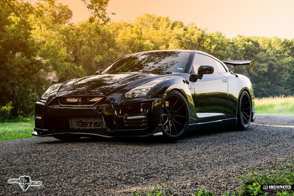 Black Nissan GT-R with aftermarket wheels and carbon fiber aero kit parked on an asphalt road at sunset.