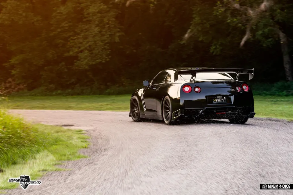 Black Nissan GT-R with a large aftermarket wing and custom wheels driving on a tree-lined road at sunset.