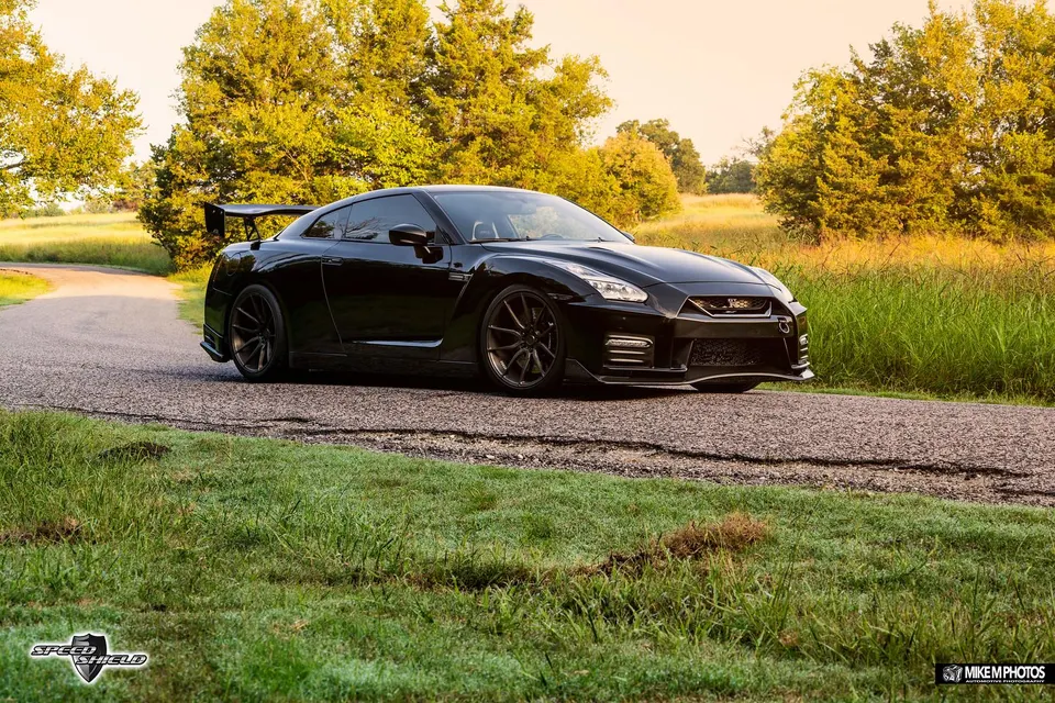 A black Nissan GT-R R35 with aftermarket wheels and a large rear wing parked on a road with trees in the background.
