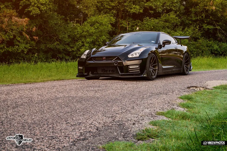 Black Nissan GT-R with aftermarket wheels and a large rear wing parked on a paved road with green trees.
