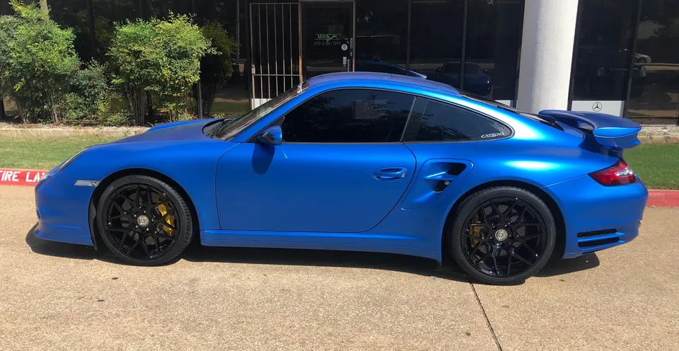 A blue Porsche 911 Turbo with black wheels and yellow brake calipers parked outside a building.