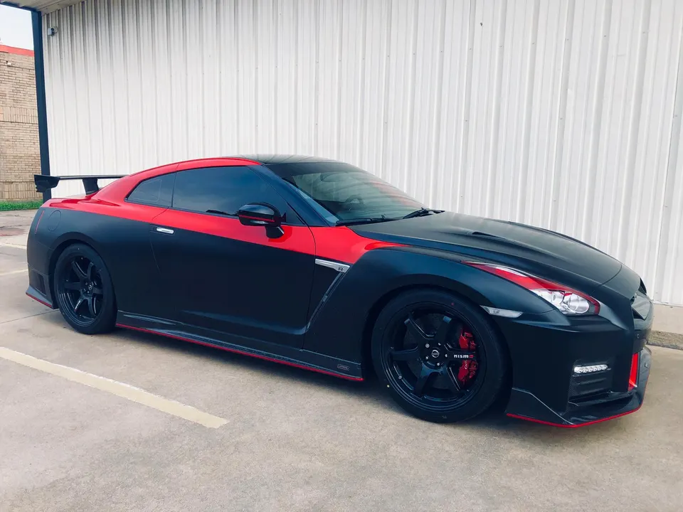 A two-tone matte black and red Nissan GT-R R35 with aftermarket wheels and a rear wing parked against a white wall.