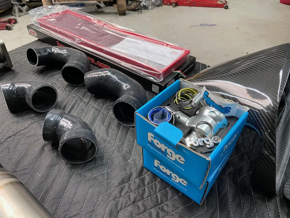 Performance car parts laid out for installation, including a BMC air filter, Forge Motorsport blow-off valve, silicone hoses, and carbon fiber components.