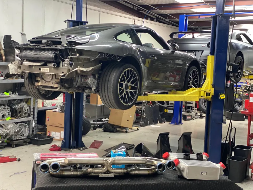 A grey Porsche 911 on a lift in a performance tuning shop, with aftermarket exhaust and intercooler parts on a table.