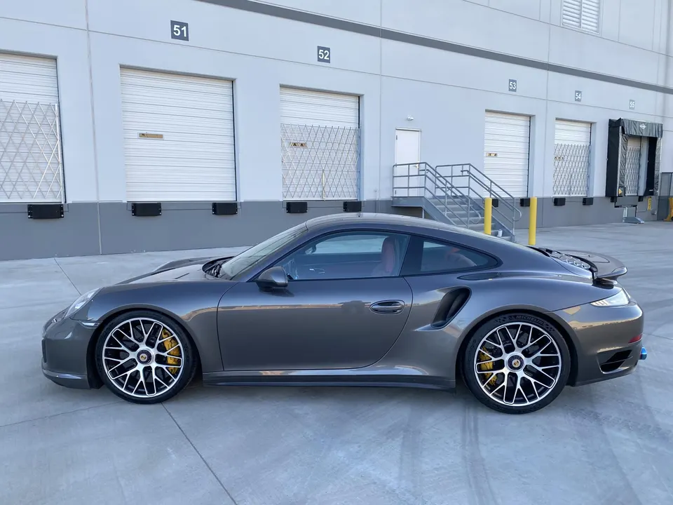 A grey Porsche 911 Turbo S parked outside a warehouse building, showcasing its side profile and yellow brake calipers.