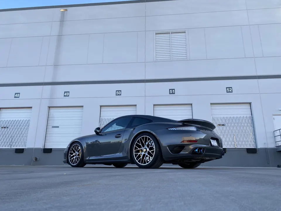 A grey Porsche 911 with aftermarket multi-spoke wheels and blue exhaust tips parked in front of an industrial building.