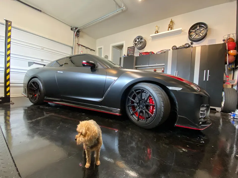 Matte black Nissan GT-R with red accents and aftermarket wheels parked in a clean garage with a small dog.