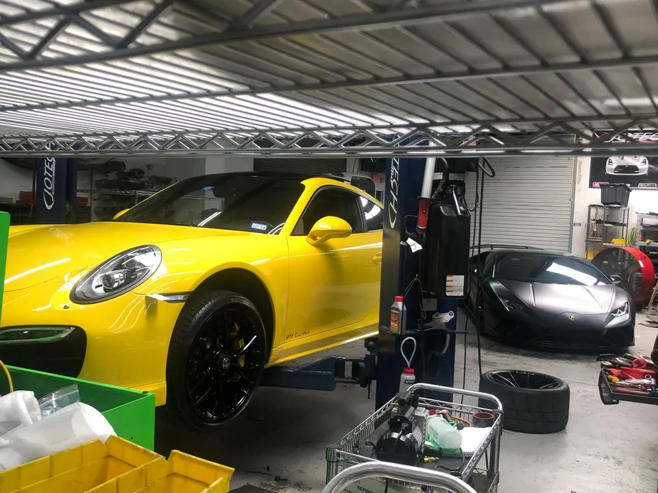 A yellow Porsche 911 Turbo on a lift in a performance tuning shop with a black Lamborghini Huracan parked nearby.