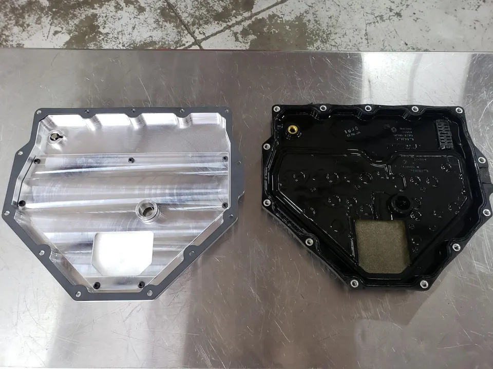 Two transmission oil pans, one machined aluminum and one black OEM, on a metal workbench in a shop.