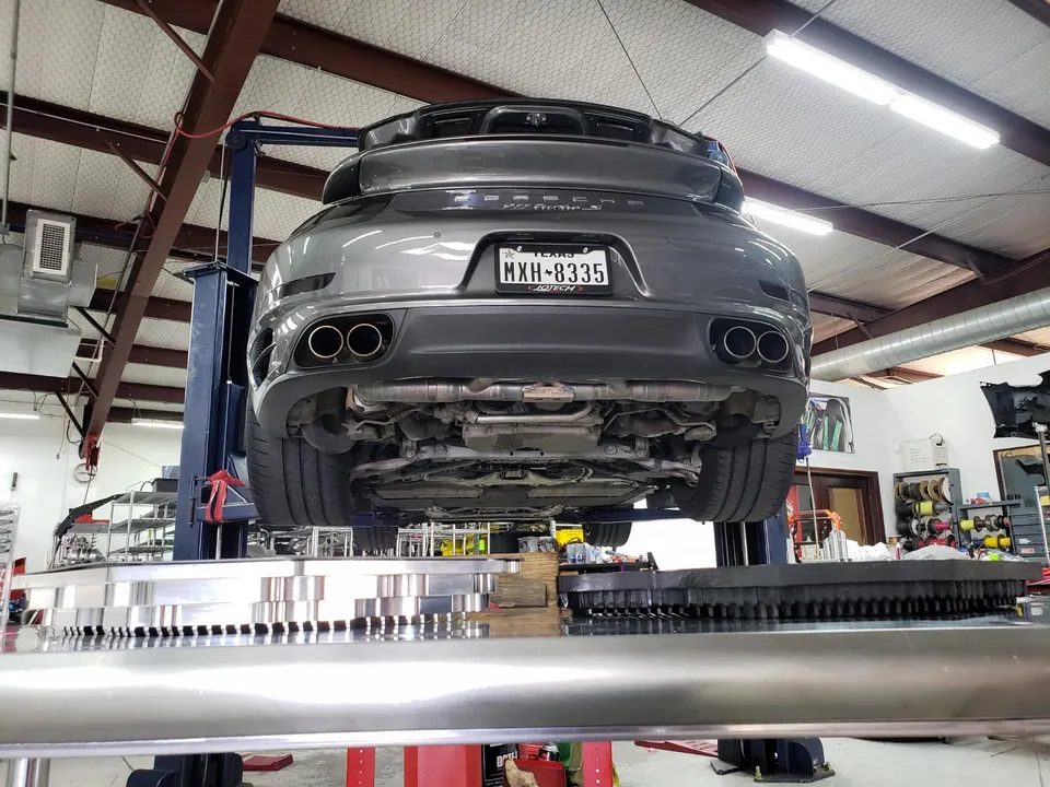 Rear view of a grey Porsche 911 Turbo S on a lift in a Jotech Motorsports shop, showcasing the exhaust system.
