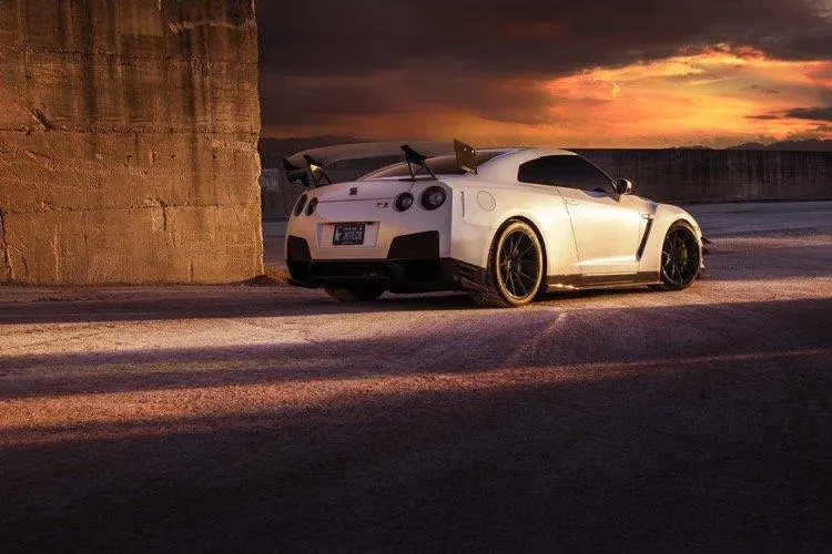 White Nissan GT-R with an aftermarket wing and diffuser parked at sunset next to a concrete wall, showcasing its exterior.