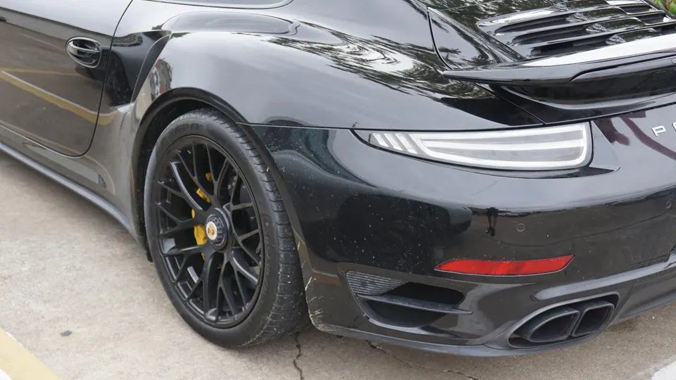 A black Porsche 911 parked on concrete, showing its rear wheel with yellow brake calipers and dual exhaust tips.