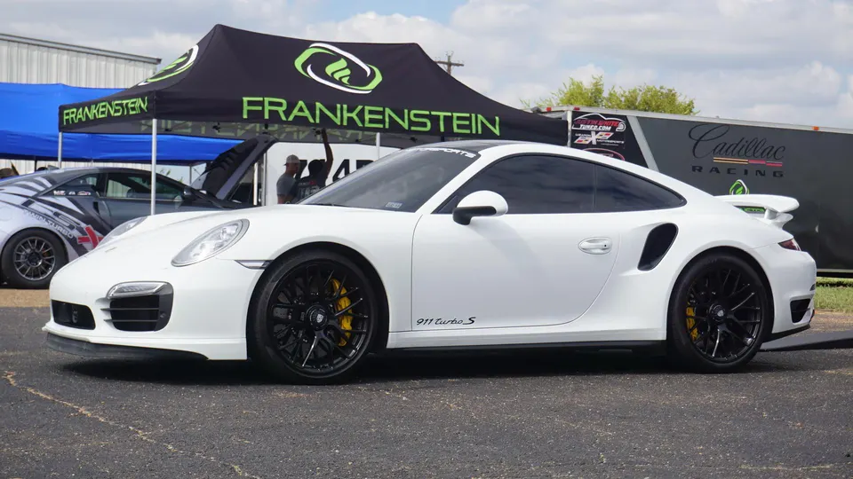 White Porsche 911 Turbo S with black wheels and yellow calipers parked at an outdoor event with Frankenstein tent in background.