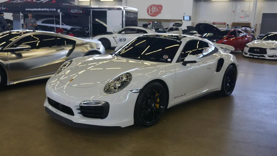 White Porsche 911 Turbo S on display at an indoor car event with other performance vehicles in the background.