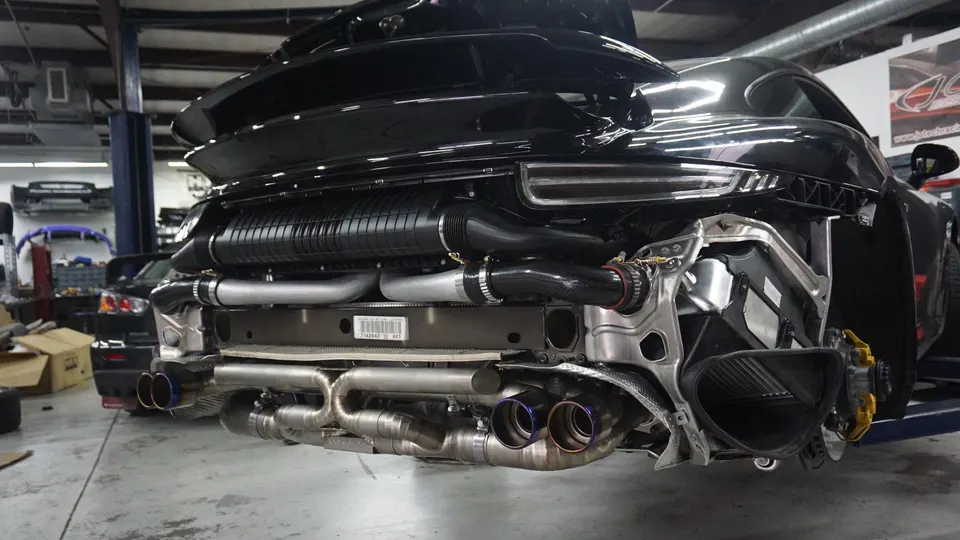 A black Porsche 911 with its rear bumper removed, showcasing a custom titanium exhaust system and intercooler piping in a performance shop.