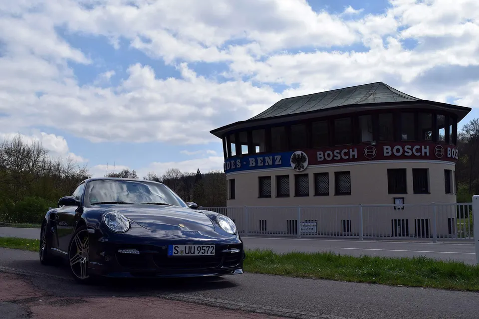 A blue Porsche 911 sports car parked on the side of a road in front of a building with Mercedes-Benz and Bosch logos.