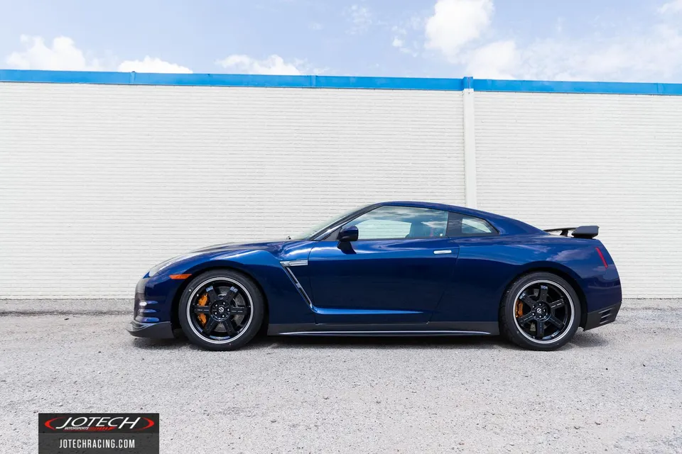 Blue Nissan GT-R parked in front of a white brick wall with a blue trim, showcasing its side profile and aftermarket wheels.