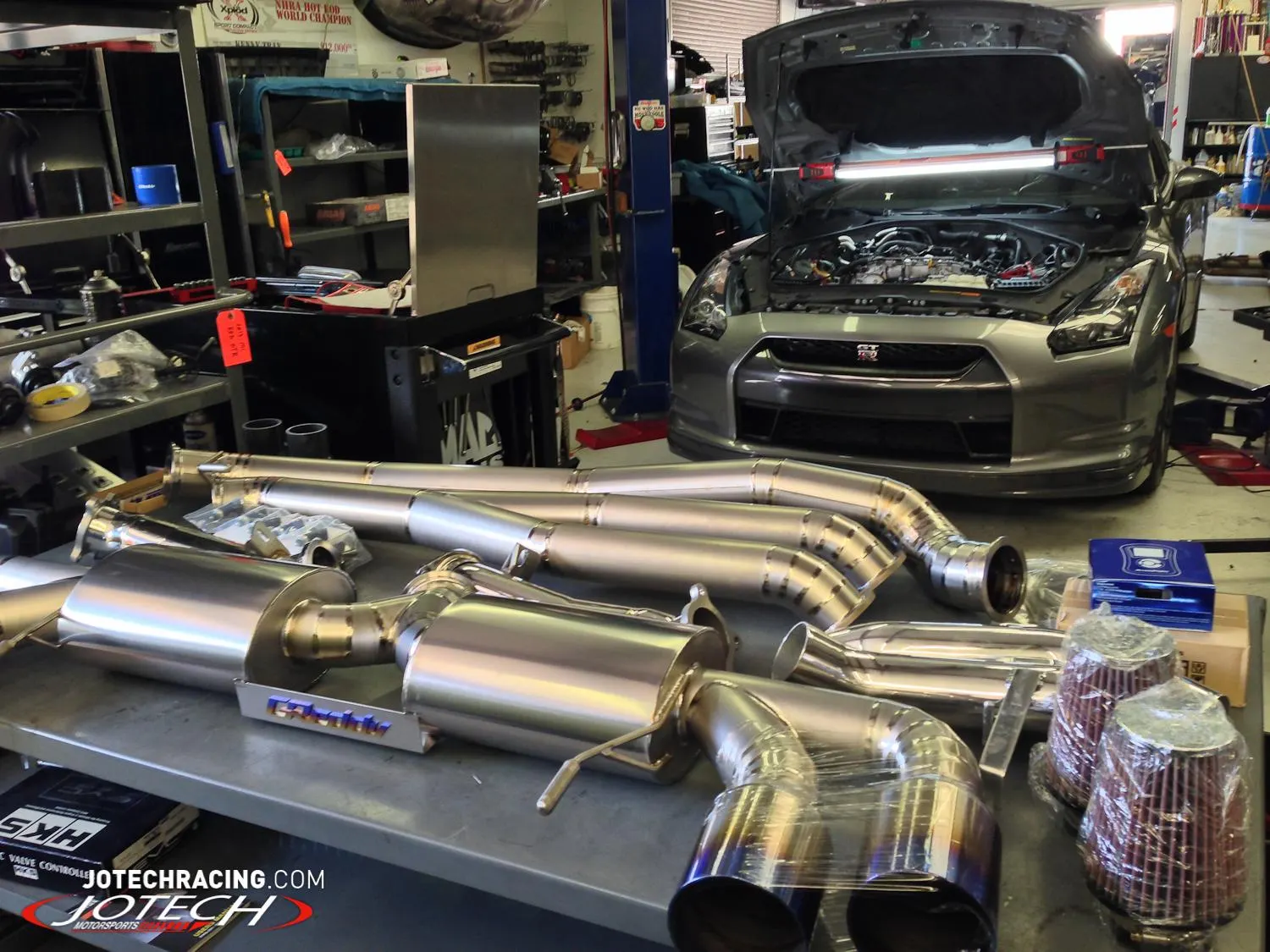 A Nissan GT-R with its hood open in a shop, with a performance exhaust system and air filters on a table.