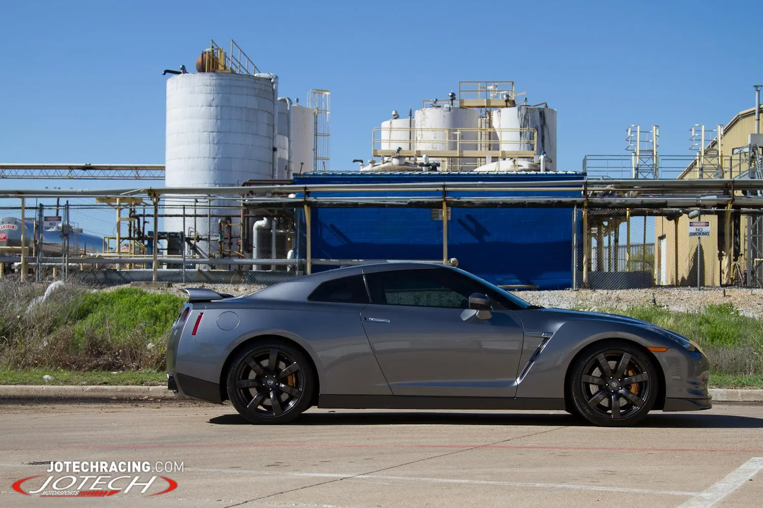 A grey Nissan GT-R R35 with black wheels parked in front of an industrial facility under a clear blue sky.