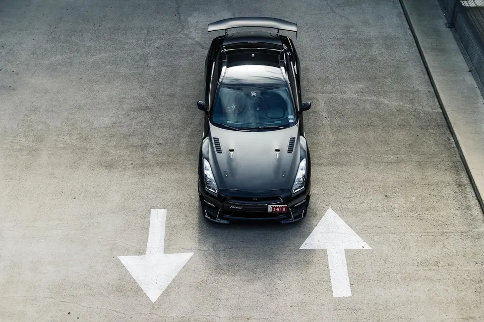 A black Nissan GT-R R35 with an aftermarket wing parked on concrete, viewed from an overhead perspective.
