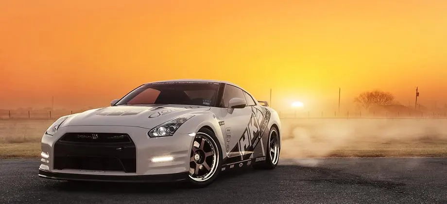 White Nissan GT-R with aftermarket wheels parked on asphalt during a vibrant sunset with mist.