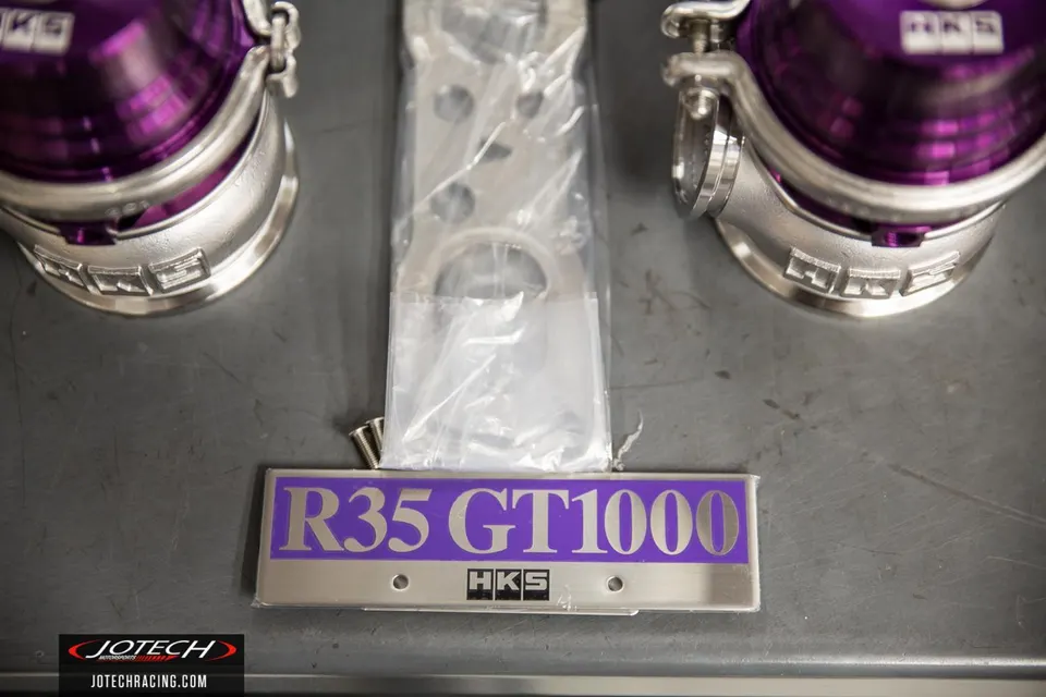 HKS R35 GT1000 blow-off valves and branding plate for a Nissan GT-R performance upgrade, laid on a dark surface.