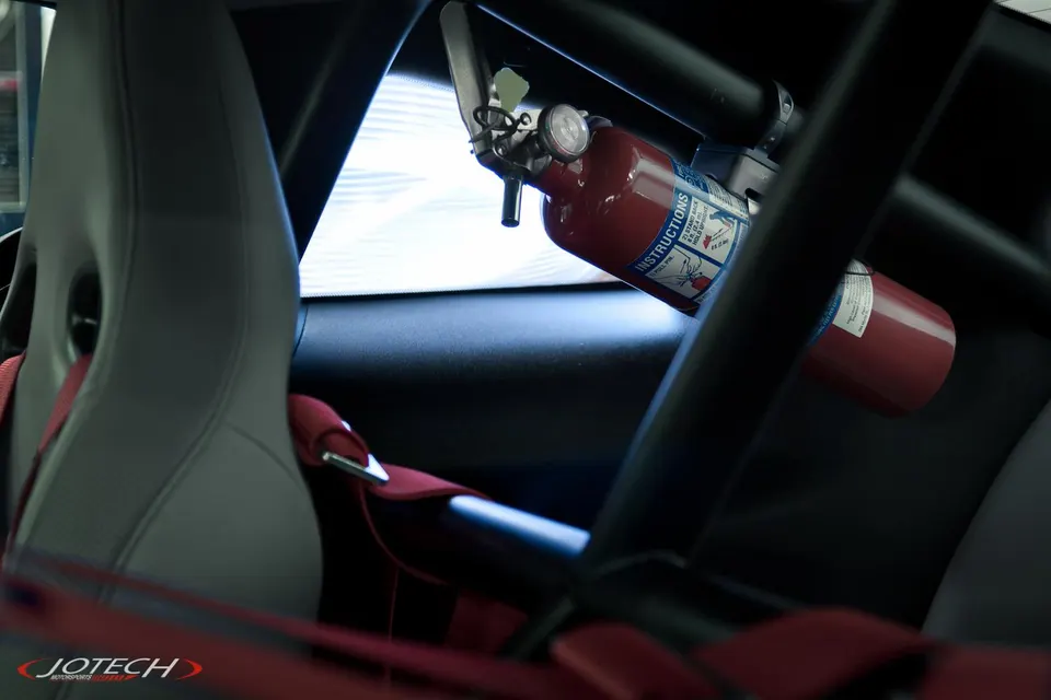 Close-up of a racing seat with red harness, roll cage, and fire extinguisher inside a performance car.