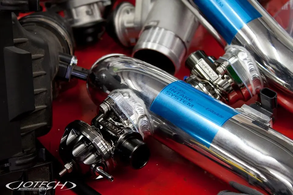 Close-up of GReddy R35 intake system components including chrome boost pipes and a blow-off valve on a red surface.