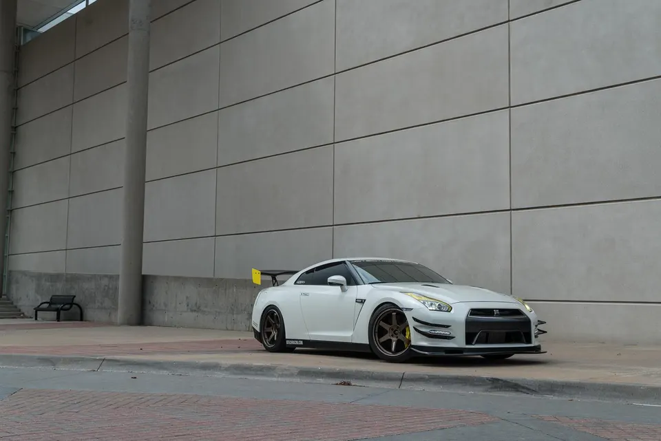 A white Nissan GT-R with aftermarket wheels and a large rear wing parked on a sidewalk next to a modern building.