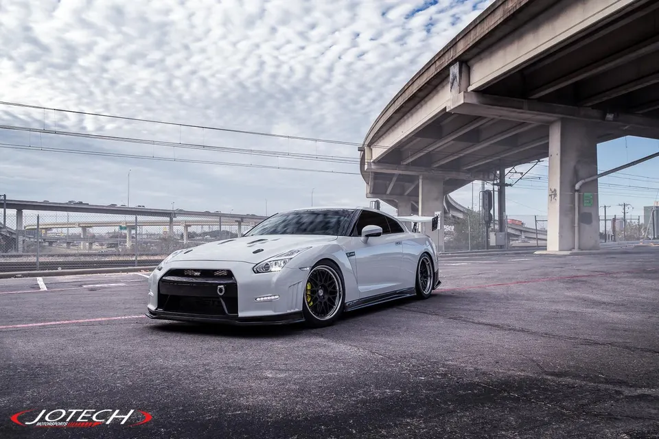 A white Nissan GT-R with aftermarket wheels and aero parked under an overpass in an urban setting.